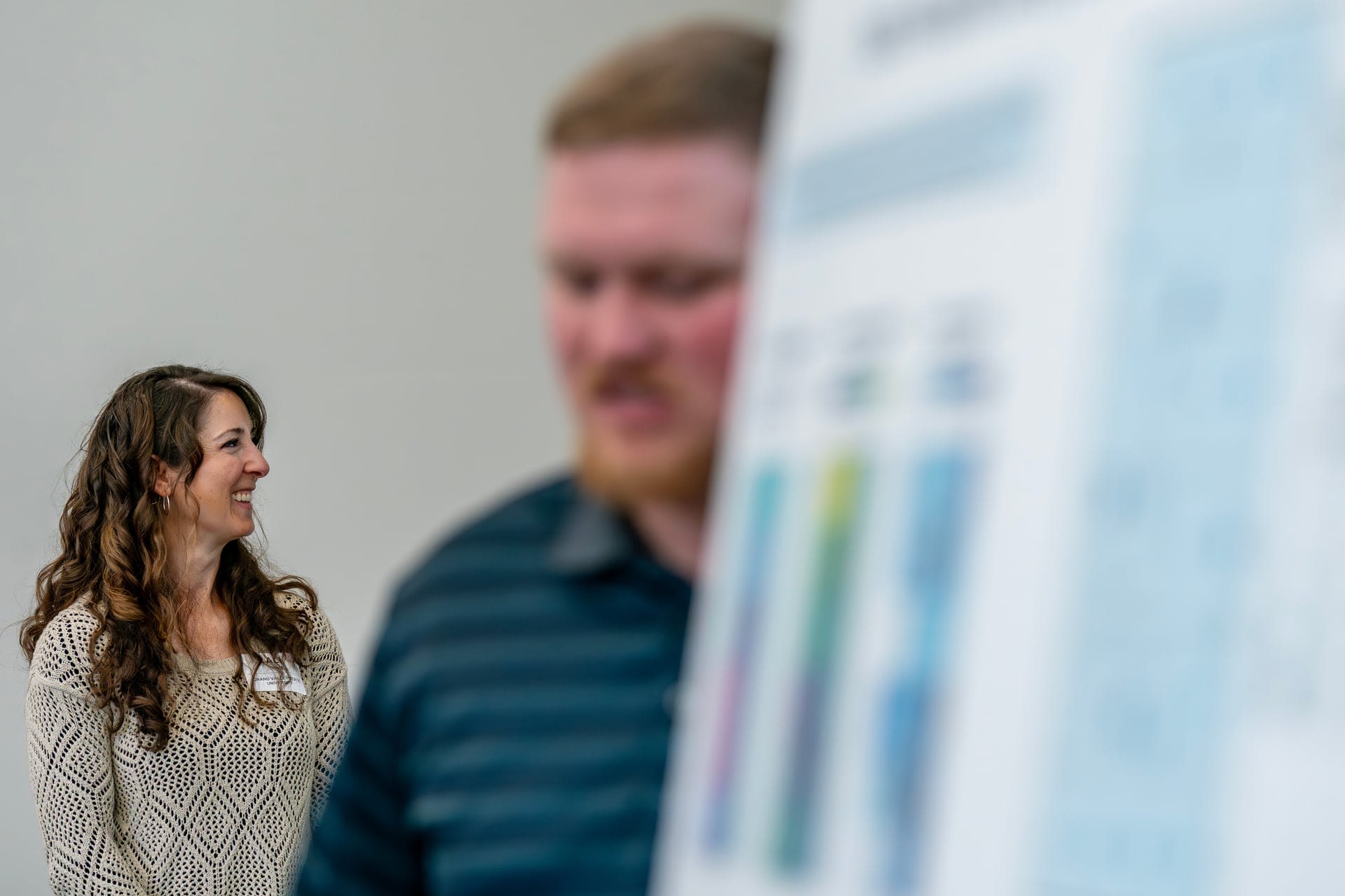 Ali Locher stands in a bright room, engaged in a conversation, while a presentation board with colorful graphics is nearby.