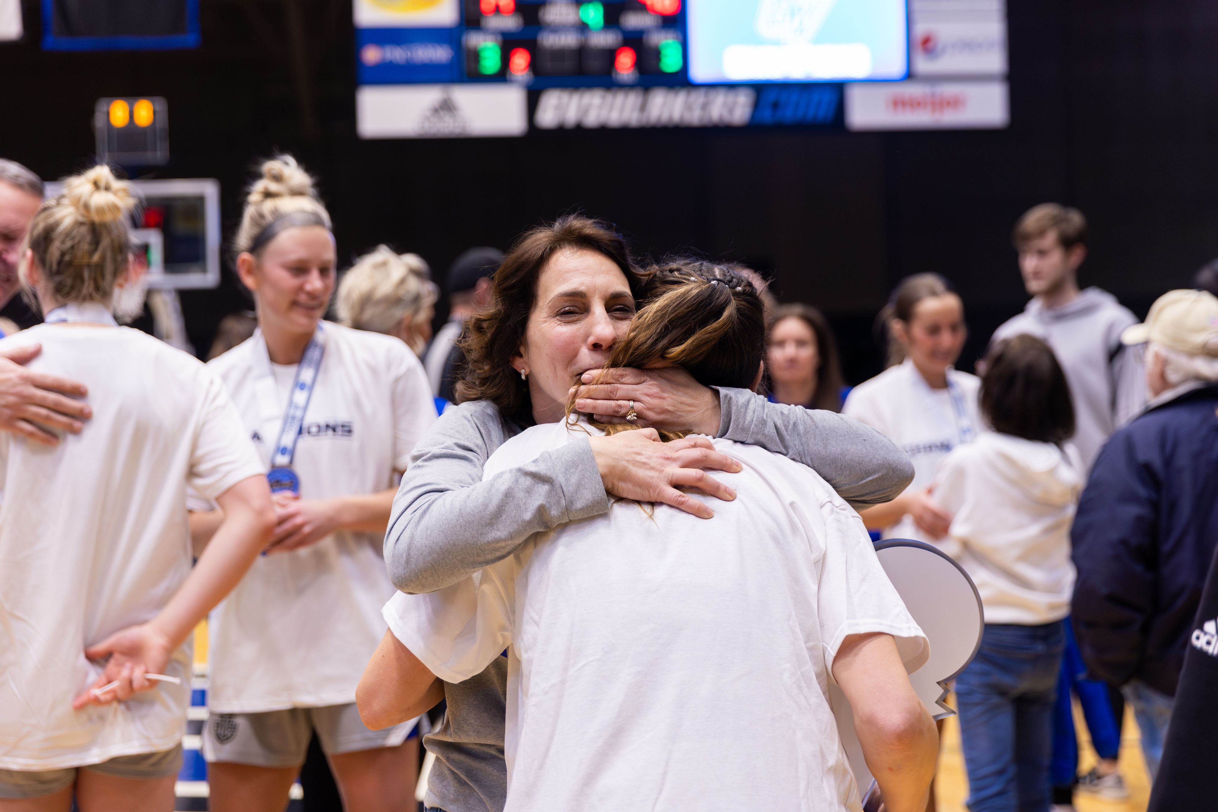 A heartfelt moment showing a woman tightly hugging a Grand Valley basketball player after a victory, with team members celebrating in the background.