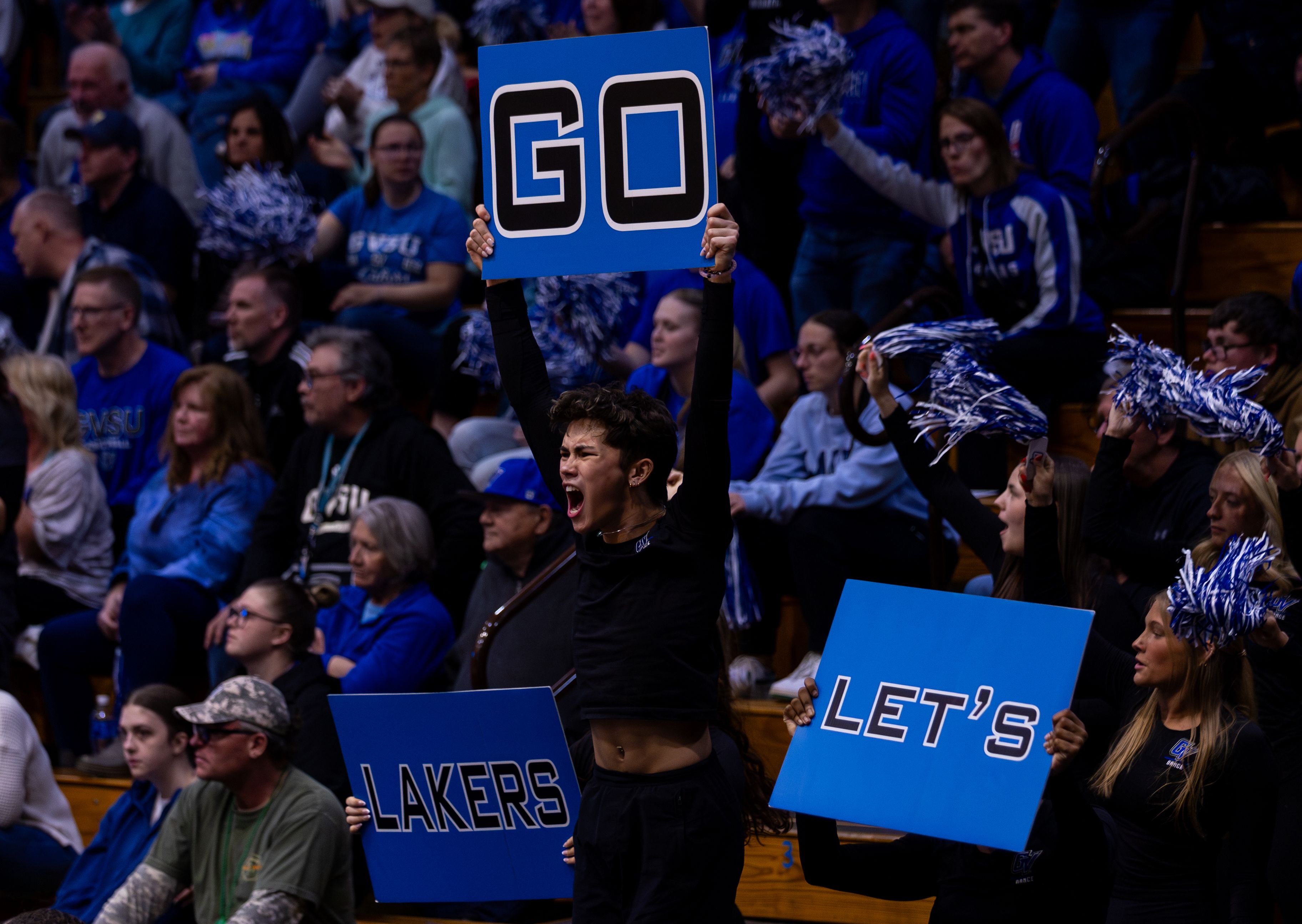 A dance member for GVSU hold's a blue sign with black letters reading 'GO' above his head as he cheers the team on in excitement.