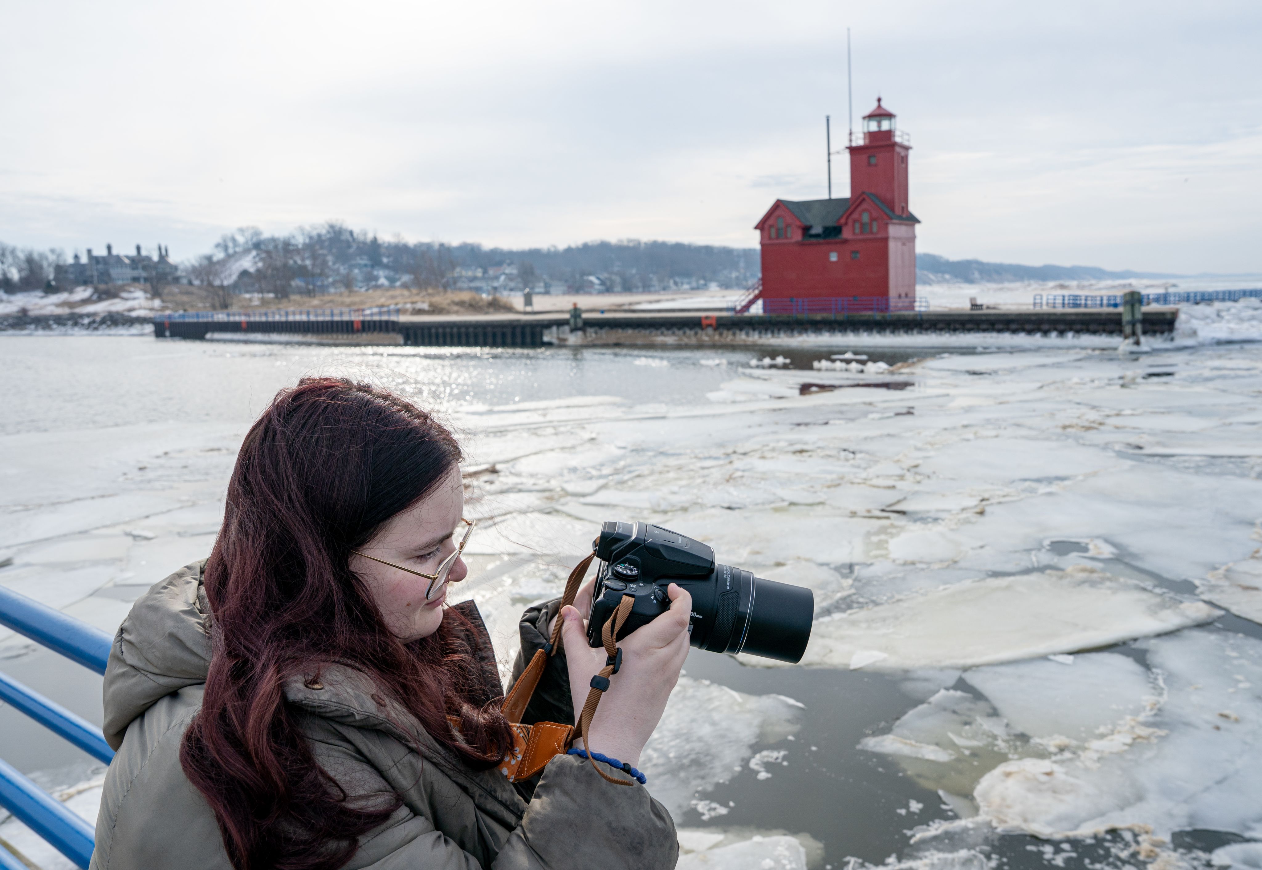 An individual holds a camera up while looking at the monitor of it. In the background sections of ice float on the water with a red building seen further back.