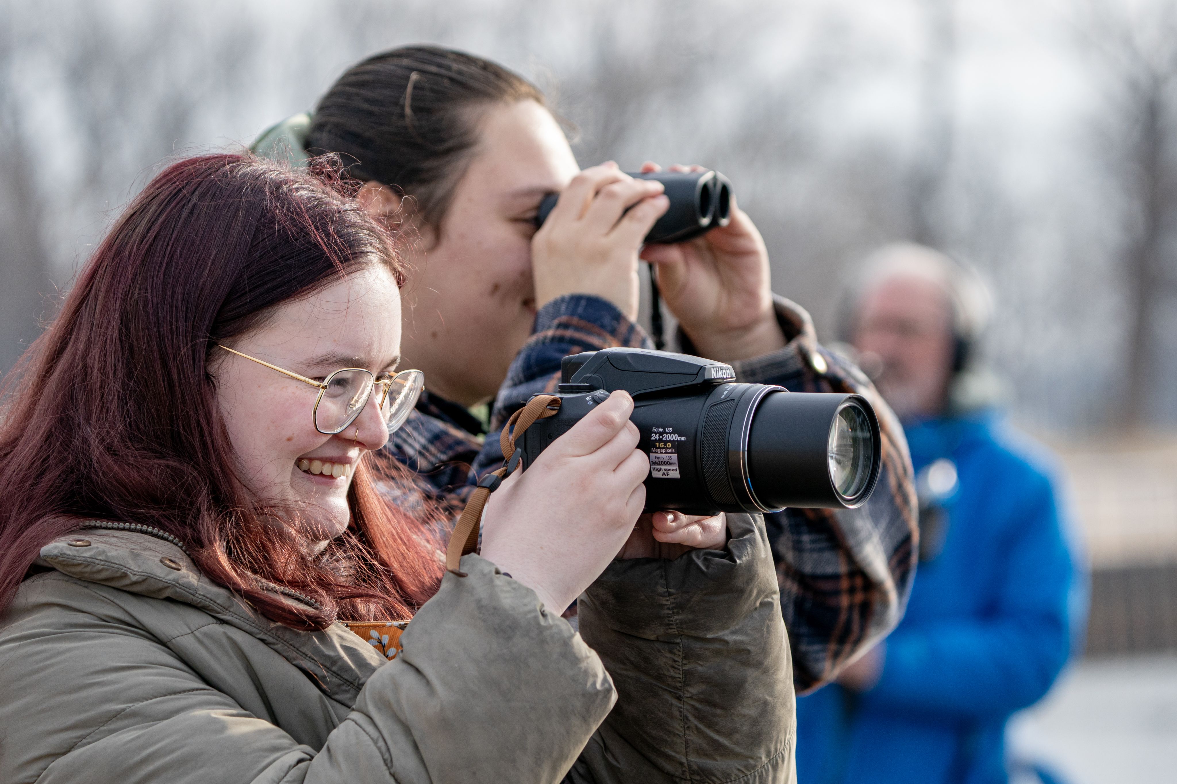 Two individuals smile while looking off to the right. One person holds a camera up, with the person behind them looking into binoculars.