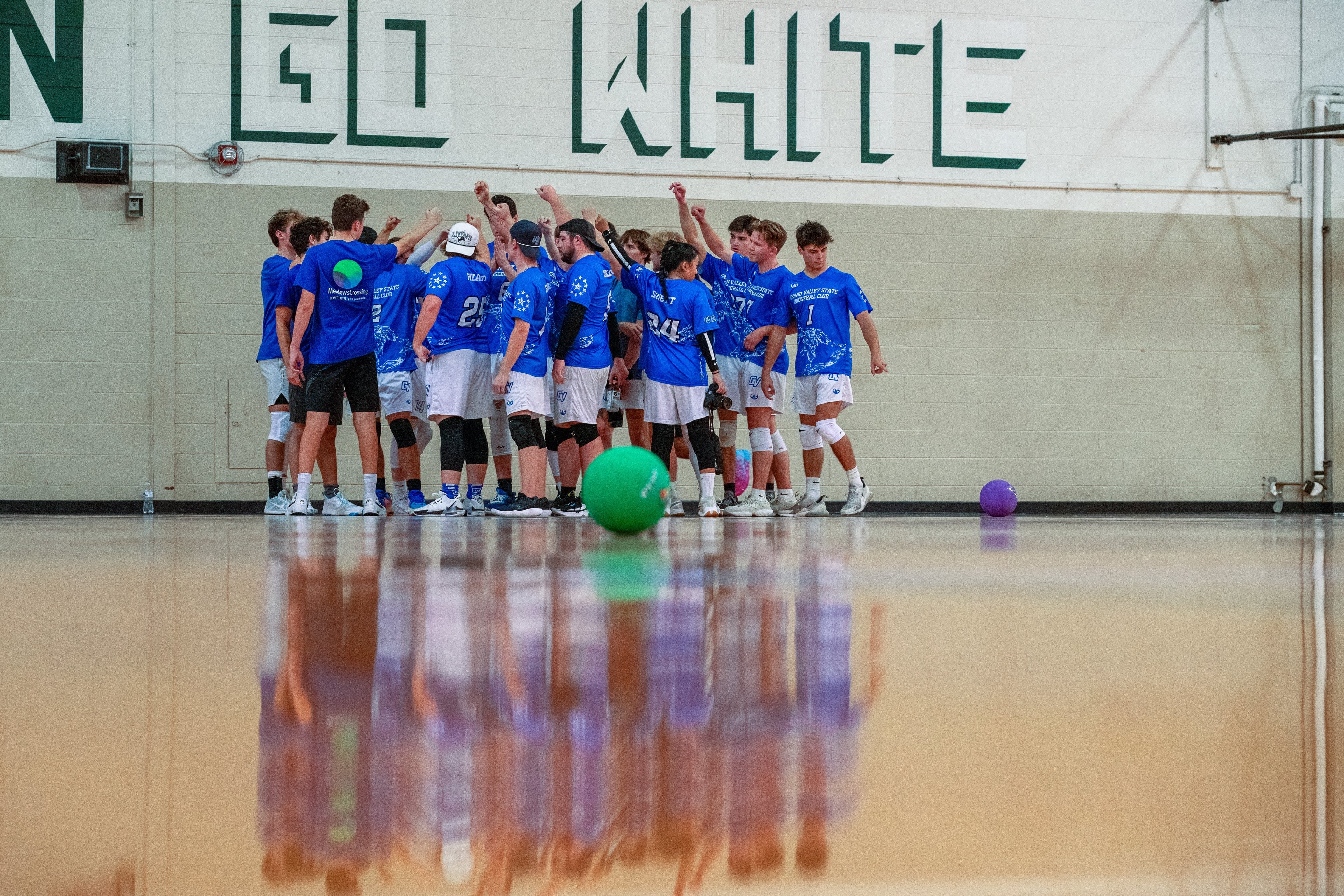 A team of dodgeball players stand on a court with their arms raised together.