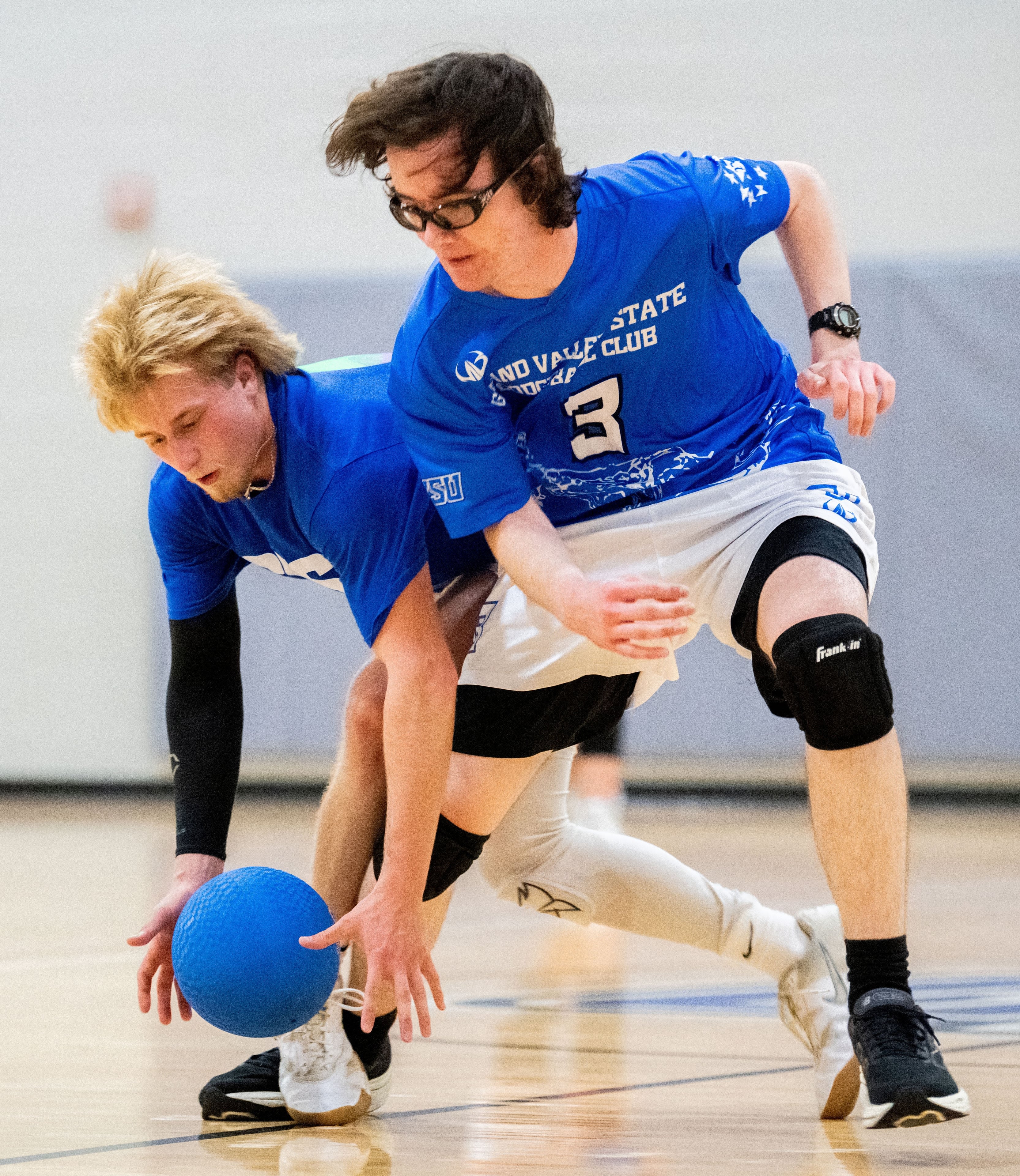Two players maneuver around each other, with one reaching down to grab a dodgeball.