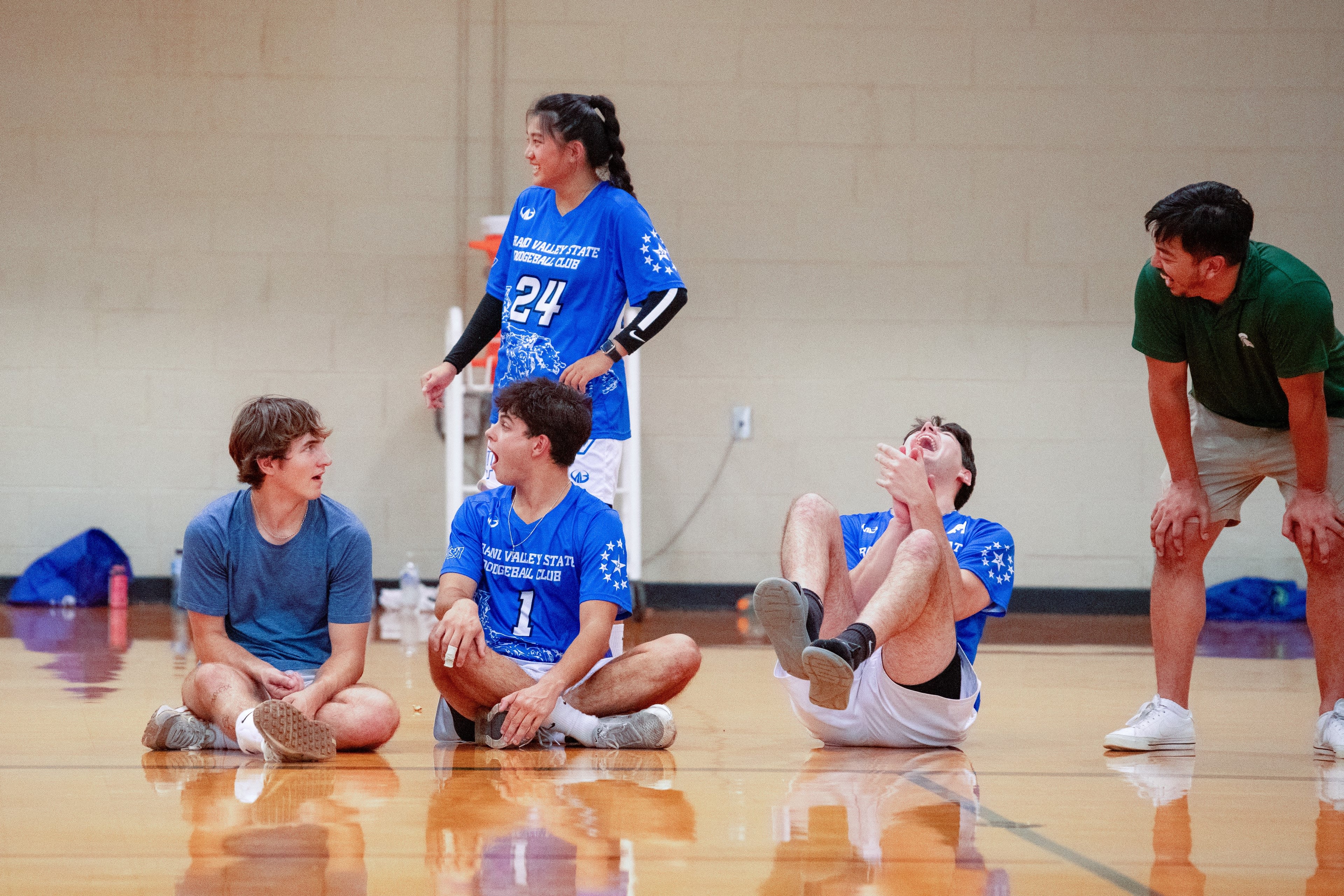Players sit and stand on the sideline reacting to the game. Two show a suprised expression, while the other three smile and laugh.