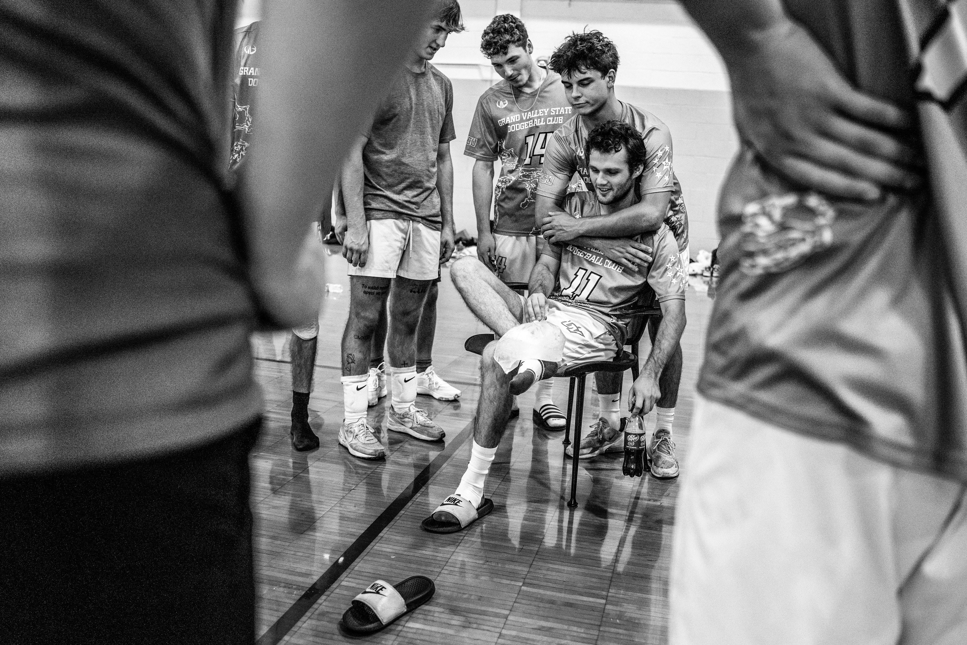 A black and white image of a player sitting down with his foot propped up to ice it as another player hugs him from behind.