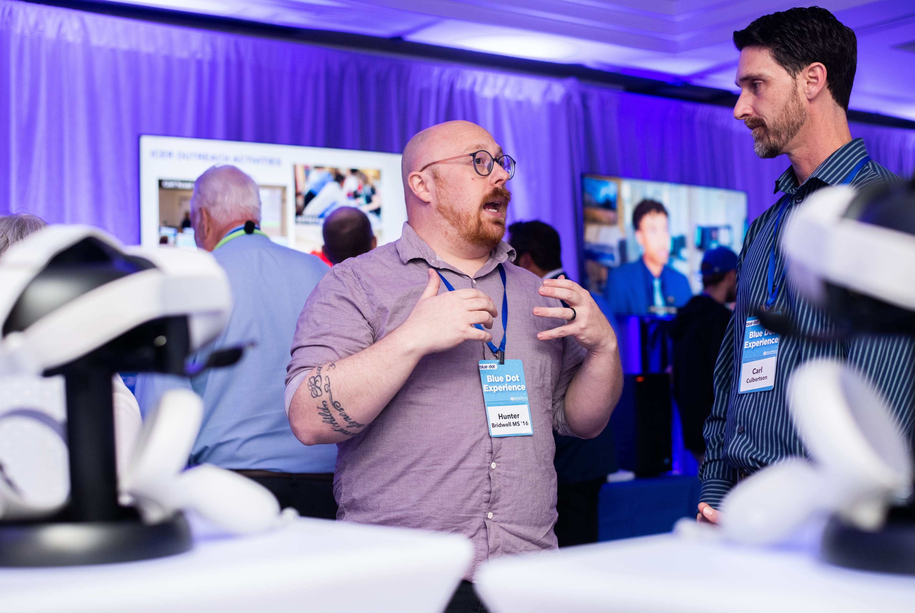 Hunter Bridwell, wearing glasses and a light purple shirt, gestures with his hands while speaking to another man at a tech event. Both wear name badges reading “Blue Dot Experience.” Virtual reality headsets are visible in the foreground, and large video screens with digital content are displayed behind them in a room lit with blue and purple lighting.