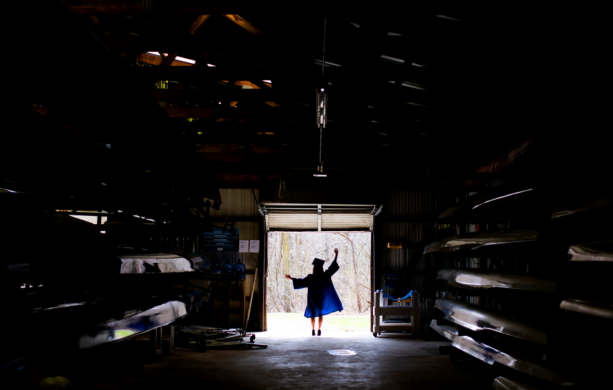 Silhouette of a person in a graduation gown and cap stands joyfully at the entrance of a dark warehouse filled with stored boats, light streaming in.