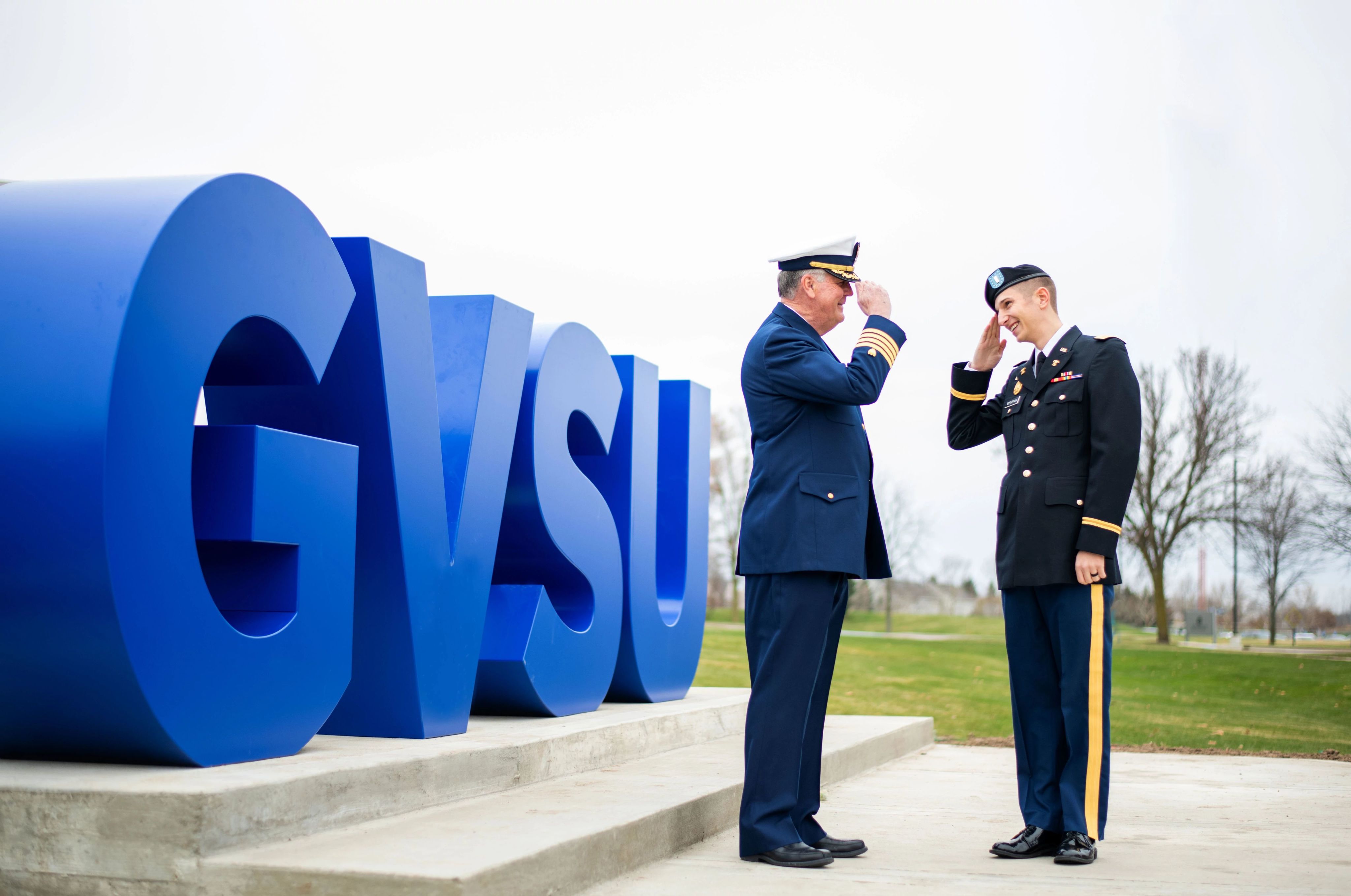 Emeritus President Tom Haas saluting a fellow veteran standing next to the big GVSU letters on campus
